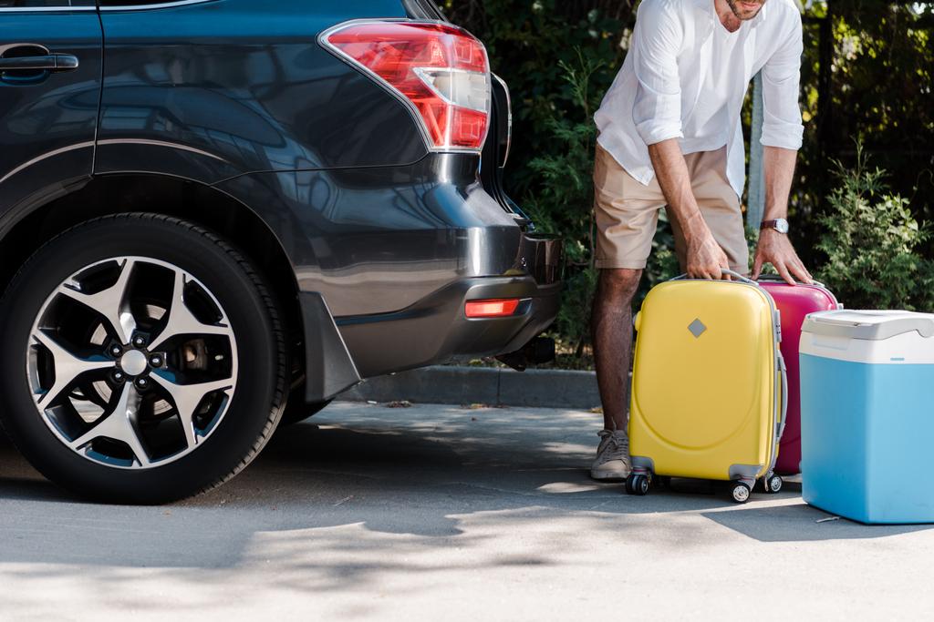 bijgesneden beeld van bebaarde man staande in de buurt van gele bagage in de buurt van auto  - Foto, afbeelding