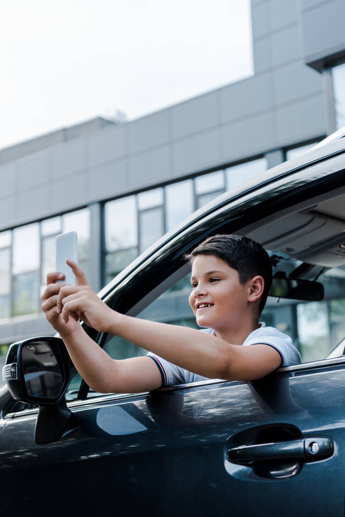 niski kąt widzenia wesoły chłopiec biorąc selfie z okna samochodu  - Zdjęcie, obraz