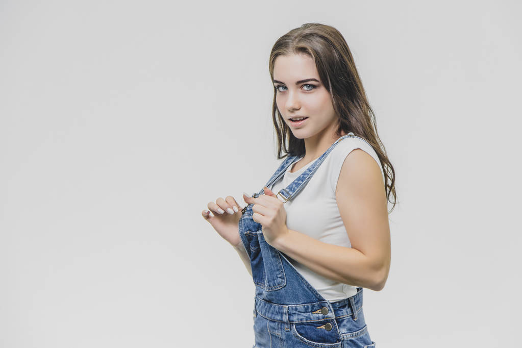 Young pretty student girl standing on a white background. Dressed in a denim suit and a white T-shirt. Holding his hands on the pliers from the combinator eyefully looks into the camera. - Photo, Image