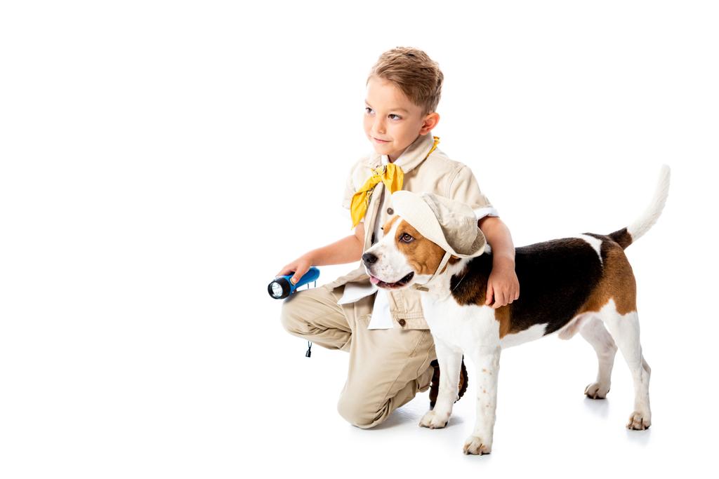 χαμογελαστός εξερευνητής παιδί αγκαλιάζοντας χαριτωμένο σκυλί κυνηγόσκυλο στο καπέλο και κρατώντας φακό σε λευκό - Φωτογραφία, εικόνα