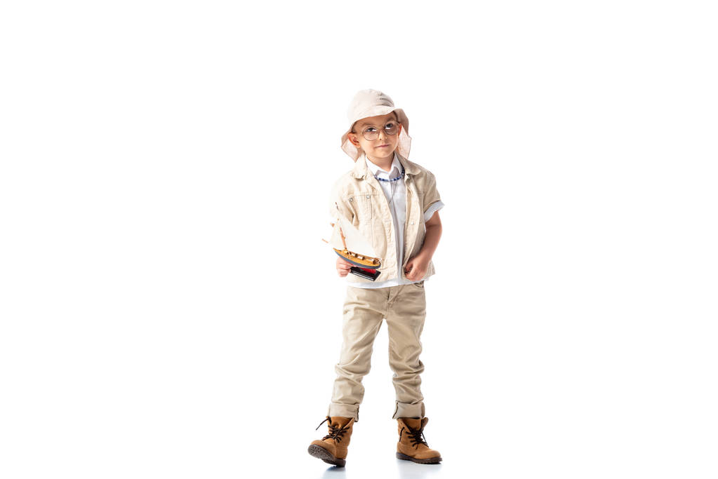 full length view of explorer child in glasses and hat holding toy ship on white - Photo, Image