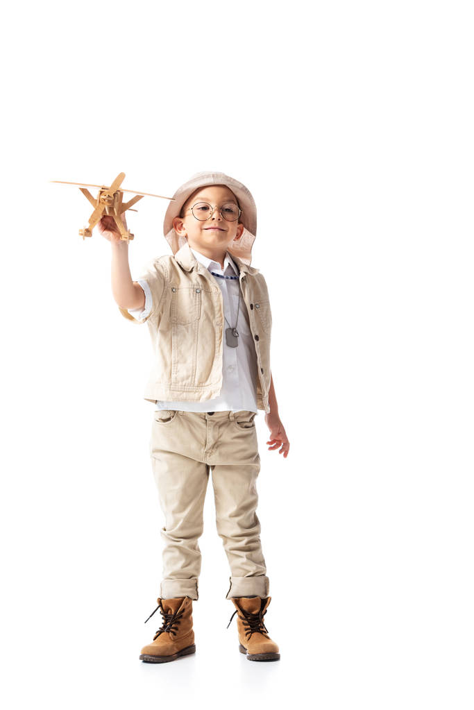 白に隔離された木製のおもちゃの飛行機を保持する眼鏡と帽子で笑顔の探検家の少年の完全な長さの眺め - 写真・画像