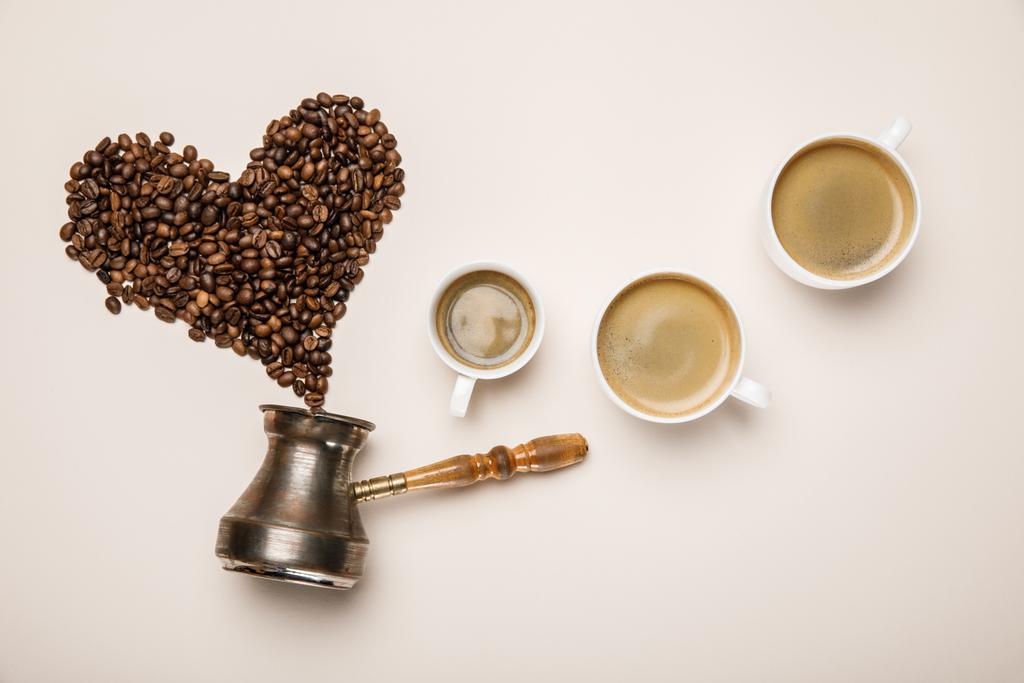top view of cups with fresh coffee near heart made of coffee grains and cezve on beige background - Photo, Image