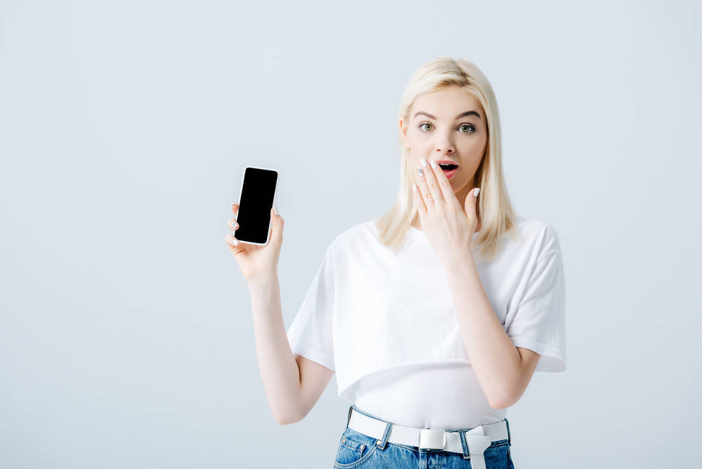 beautiful girl showing smartphone with blank screen and covering mouth isolated on grey - Photo, Image