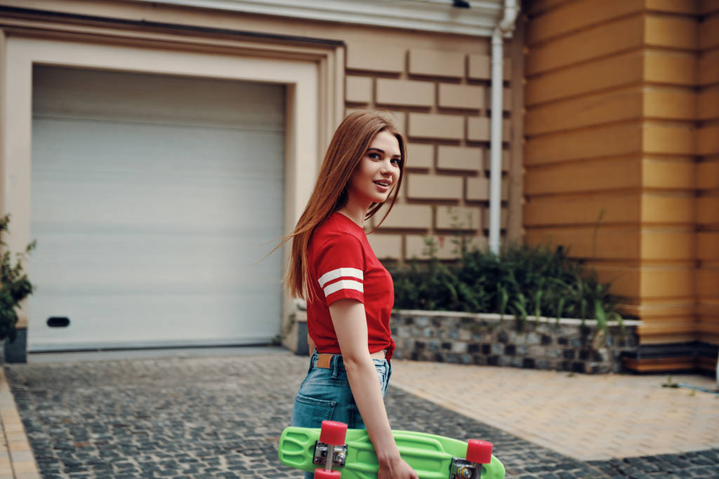 Красивая молодая женщина смотрит в камеру и носит скейтборд, стоя на улице
   - Фото, изображение