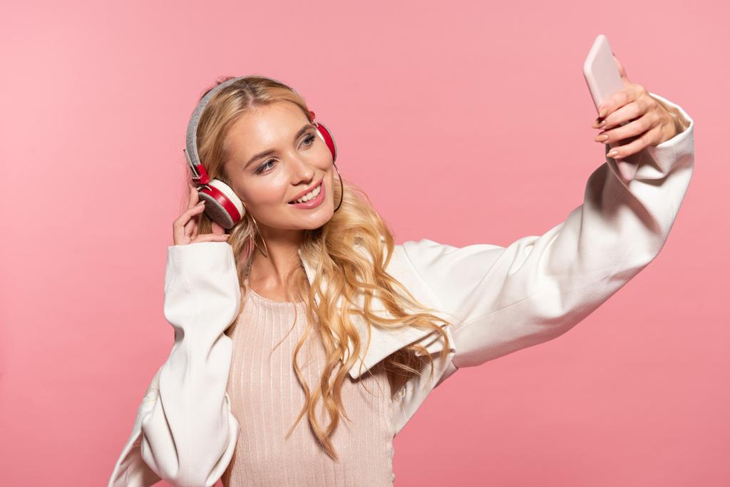 hermosa mujer rubia con auriculares tomando selfie en el teléfono inteligente aislado en rosa
 - Foto, Imagen