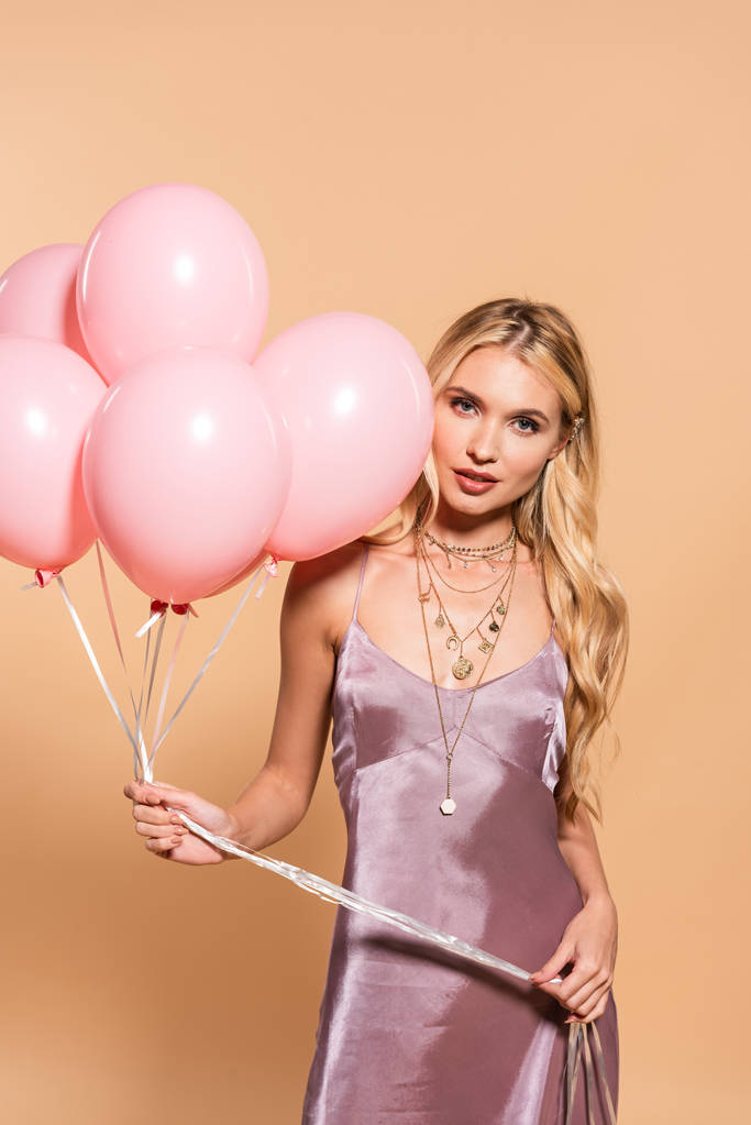 Elegancka Blondynka w fioletowej satynowej sukni i naszyjnik trzyma różowe balony na beżu - Zdjęcie, obraz