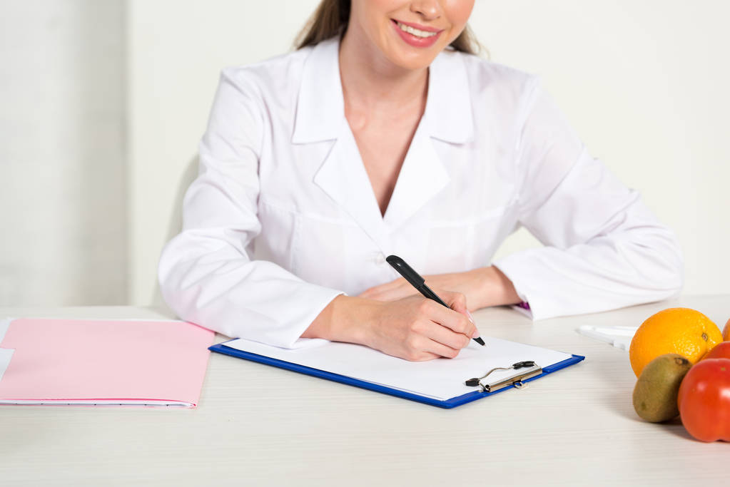 abgeschnittene Ansicht eines lächelnden Diätassistenten in weißem Mantel am Arbeitsplatz Schreiben in Klemmbrett mit Früchten auf dem Tisch - Foto, Bild