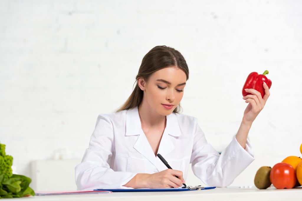 Ernährungsberaterin in weißem Mantel mit rotem Paprika und Schriftzug im Klemmbrett am Arbeitsplatz - Foto, Bild