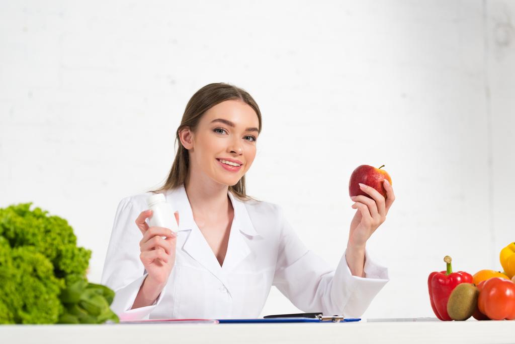улыбающийся диетолог в белом халате держит таблетки и яблоко на рабочем месте
 - Фото, изображение