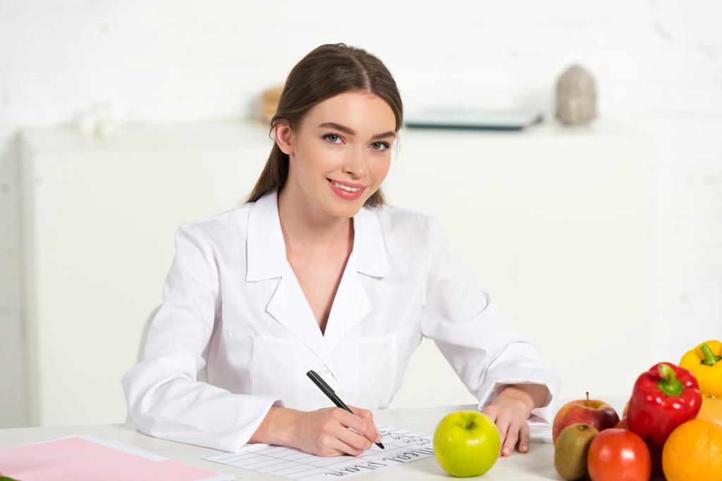 улыбающийся диетолог в белом халате пишет на рабочем месте с фруктами и овощами на столе
 - Фото, изображение