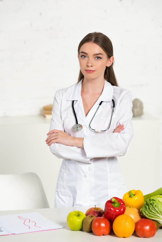 atractivo dietista en bata blanca con equipo de pie con brazos cruzados cerca de la mesa con frutas y verduras frescas
 - Foto, Imagen