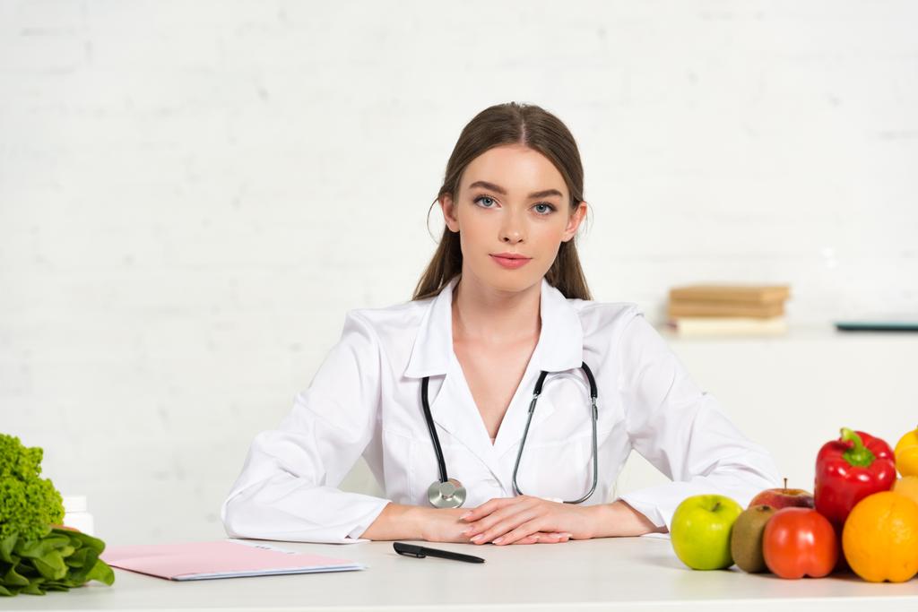 vista frontale di dietista in mantello bianco con attrezzatura seduta sul posto di lavoro con frutta e verdura sul tavolo
 - Foto, immagini