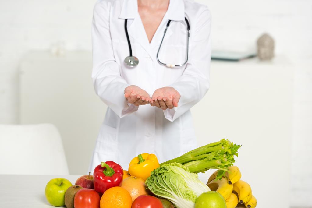 新鮮な果物や野菜の近くに聴診器を持つ白いコートの栄養士の部分的なビュー - 写真・画像