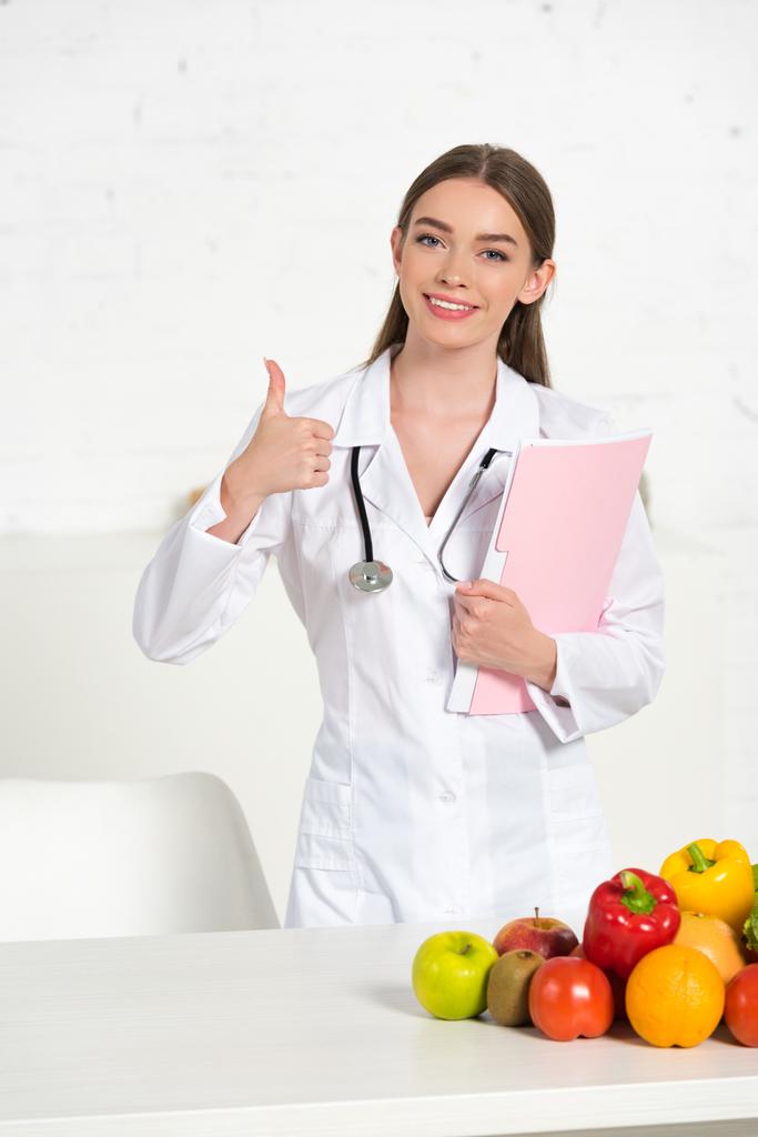 uśmiechnięty dietetyk w białym płaszczu gospodarstwa folder i pokazując kciuk w pobliżu stołu ze świeżych owoców i warzyw - Zdjęcie, obraz