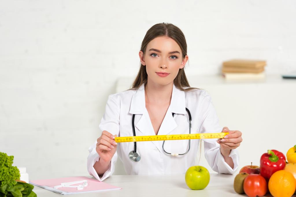 front view of dietitian in white coat with stethoscope holding measure tape at table with fruits and vegetables - Photo, Image