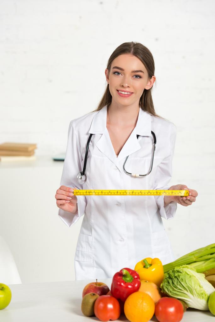 front view of smiling dietitian in white coat with stethoscope holding measure tape  - Photo, Image