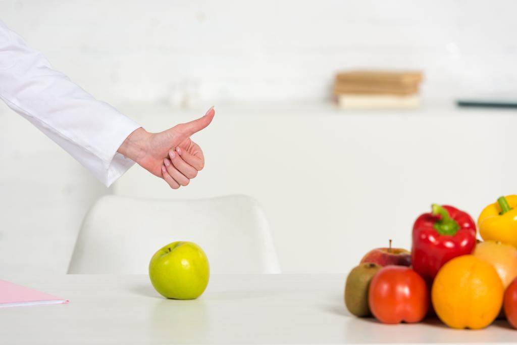 cropped view of dietitian showing thumb up near table with fresh vegetables and fruits - Photo, Image