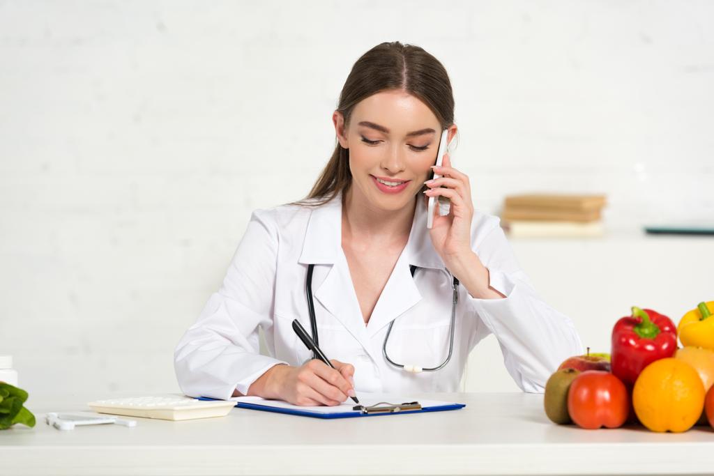 hymyilevä ravitsemusteraani valkoisessa takissa puhuminen älypuhelimella ja kirjoittaminen leikepöydällä työpaikalla
 - Valokuva, kuva