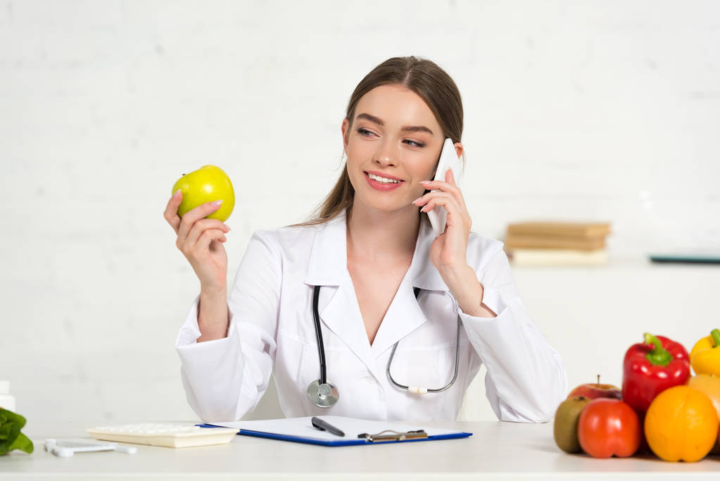 nutricionista sorridente em casaco branco falando no smartphone e segurando maçã no local de trabalho
 - Foto, Imagem