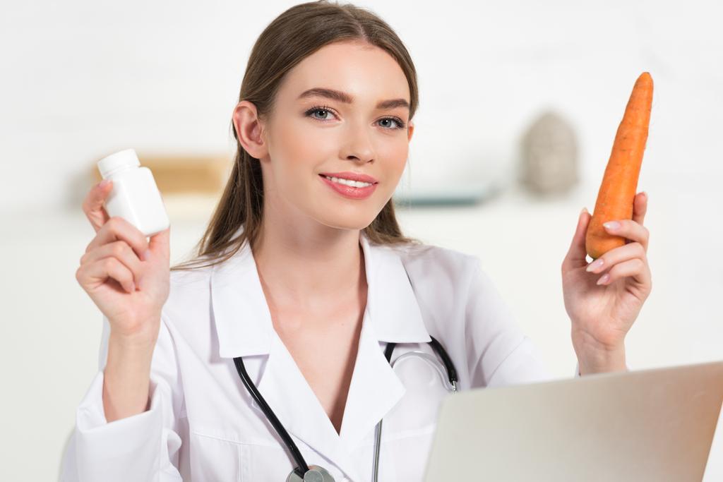dietista sonriente con abrigo blanco sosteniendo pastillas y zanahoria cerca de la computadora portátil
 - Foto, imagen