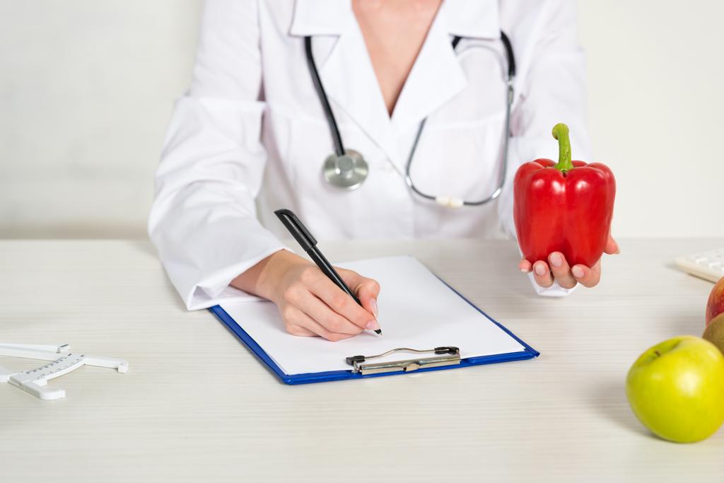 cropped view of dietitian in white coat writing in clipboard and holding bell pepper at workplace - Photo, Image