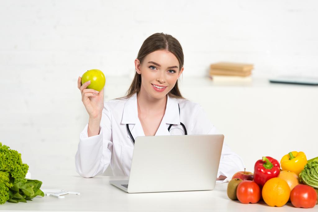 nutricionista sorridente em casaco branco segurando maçã no local de trabalho com laptop
 - Foto, Imagem