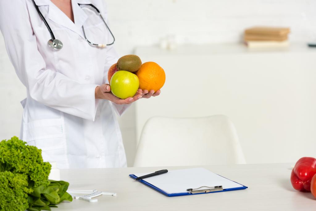 vista parcial del dietista en bata blanca sosteniendo frutas frescas cerca del lugar de trabajo
 - Foto, imagen