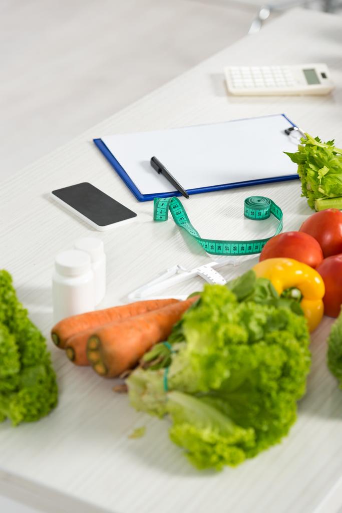 Буфер обмена с ручкой, измерительная лента, смартфон с чистым экраном, калькулятор, лекарства и свежие овощи на столе
 - Фото, изображение