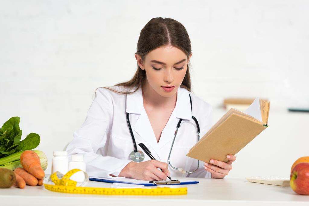 keskittynyt ravitsemusterapeutin valkoinen takki käsittelyssä kirja ja kirjallisesti leikepöydällä työpaikalla
 - Valokuva, kuva