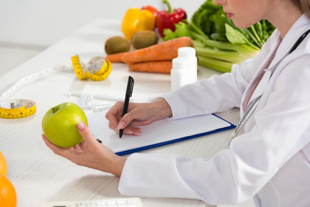 Ausgeschnittene Ansicht von Diätassistentin im weißen Mantel mit grünem Apfel und Schrift im Klemmbrett - Foto, Bild