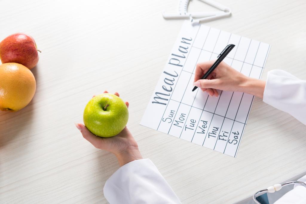 Ausgeschnittene Ansicht einer Ernährungsberaterin, die am Arbeitsplatz Apfel in der Hand hält und in den Speiseplan schreibt - Foto, Bild