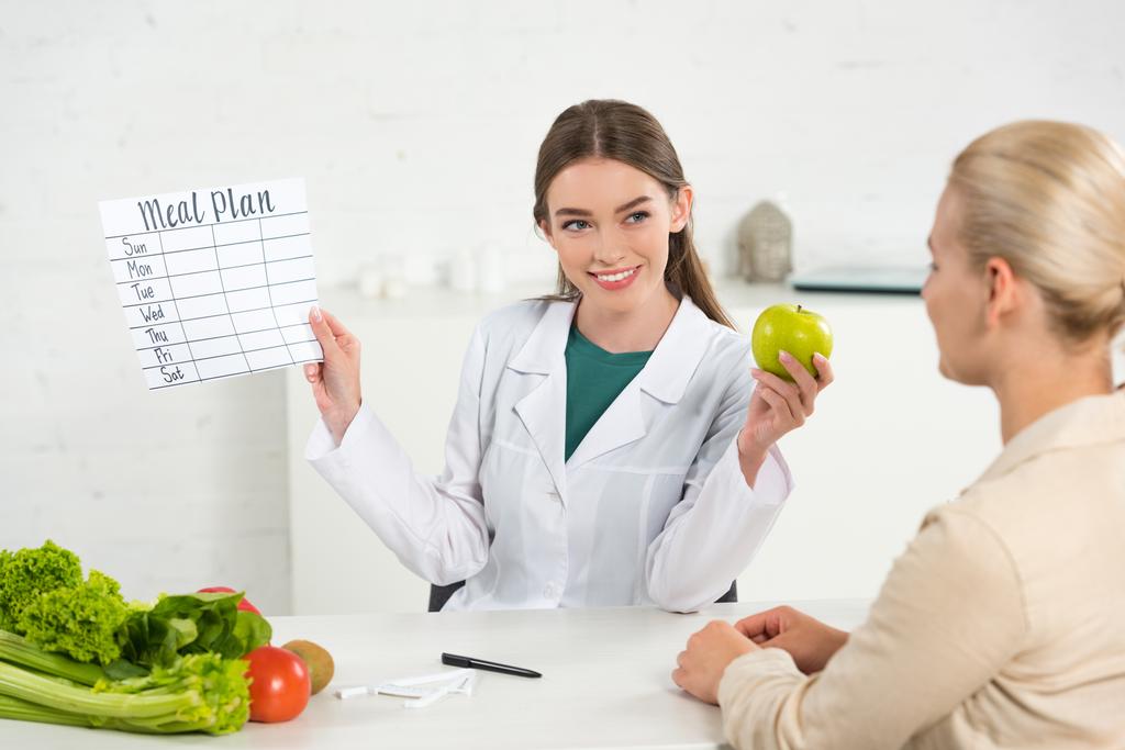 χαμογελαστός διαιτολόγος σε λευκό παλτό κρατώντας σχέδιο φαγητού και μήλο και ασθενή στο τραπέζι - Φωτογραφία, εικόνα