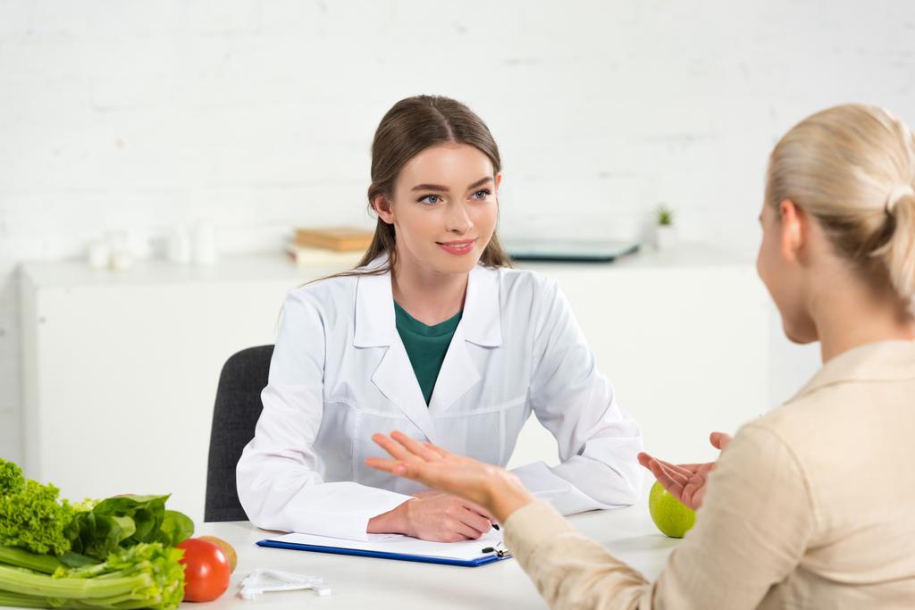 χαμογελαστός διαιτολόγος σε λευκό παλτό κοιτάζοντας τον ασθενή στο τραπέζι - Φωτογραφία, εικόνα