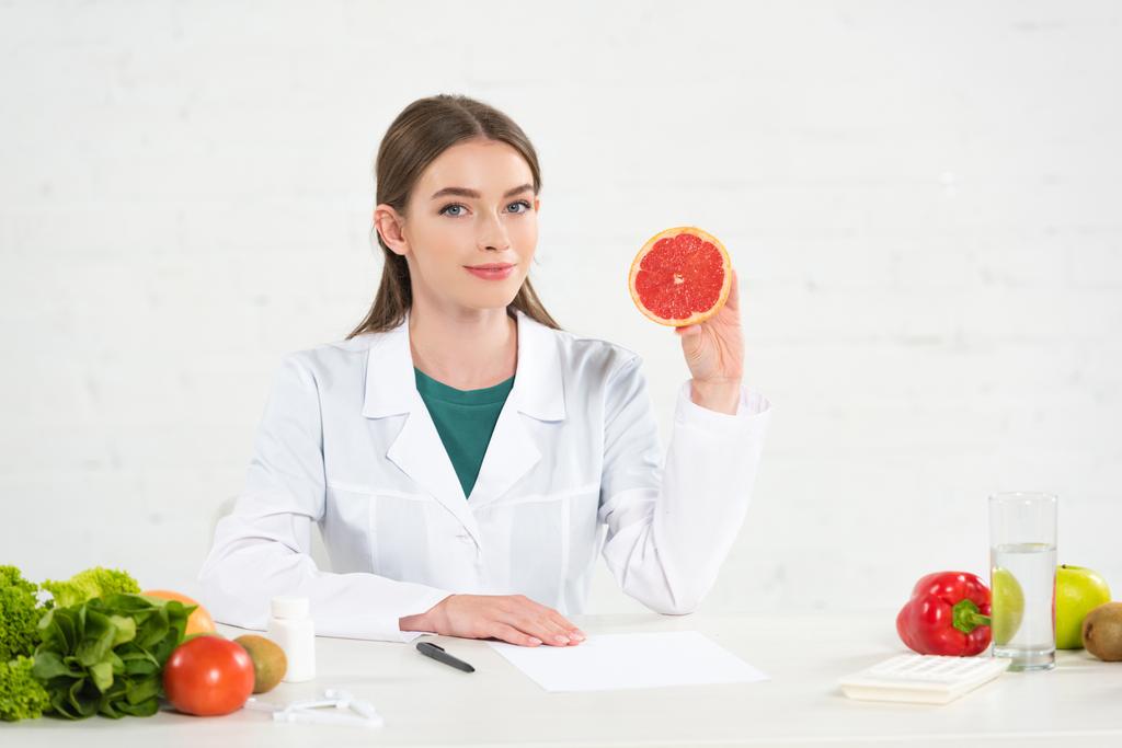 dietista en bata blanca sosteniendo toronja cortada en el lugar de trabajo
 - Foto, imagen