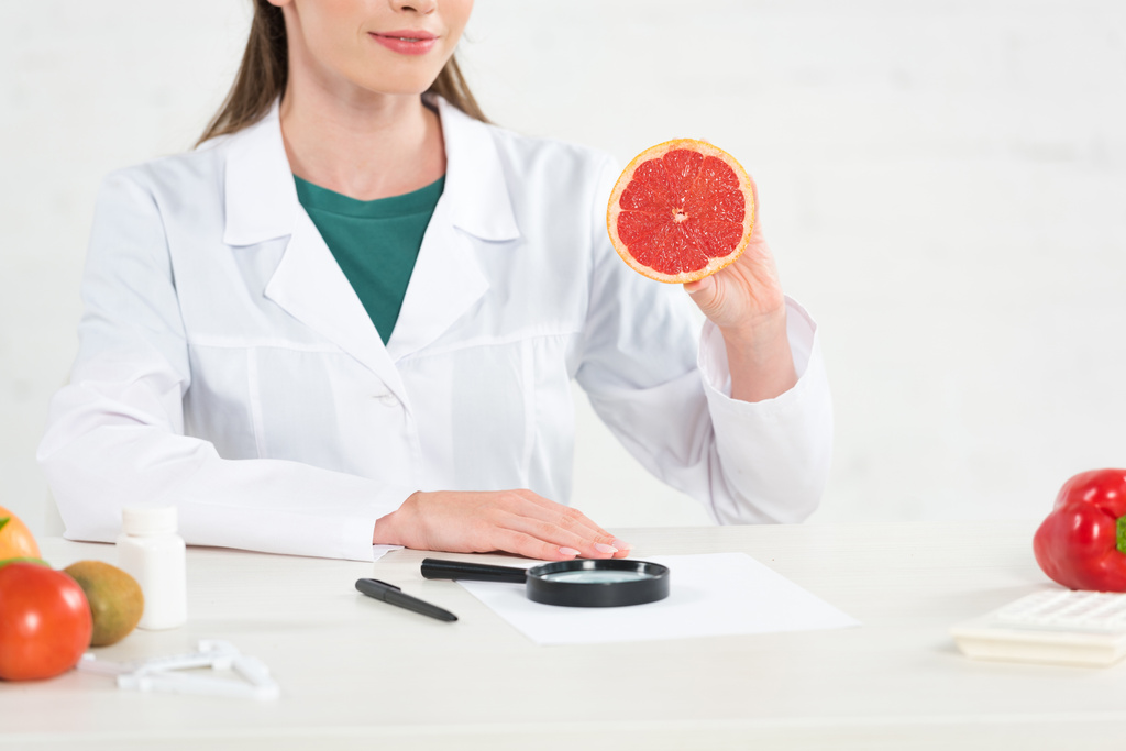 vista recortada de dietista en capa blanca sosteniendo toronja cortada en el lugar de trabajo
 - Foto, Imagen