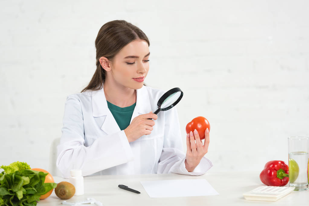 Diätassistentin im weißen Kittel betrachtet Tomate durch Vergrößerungsglas - Foto, Bild