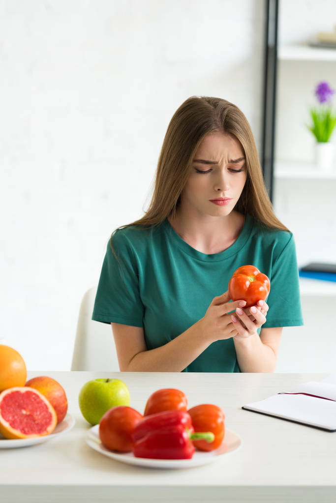 грустная женщина сидит за столом с фруктами и овощами и держит помидор
 - Фото, изображение