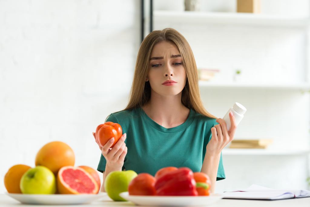 femme triste assis à table avec des fruits et légumes et tenant des pilules et de la tomate
 - Photo, image