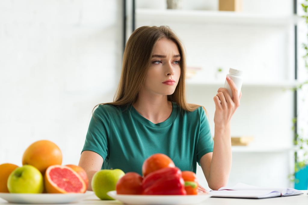 smutna kobieta siedzi przy stole z owocami i warzywami i trzyma pigułki - Zdjęcie, obraz