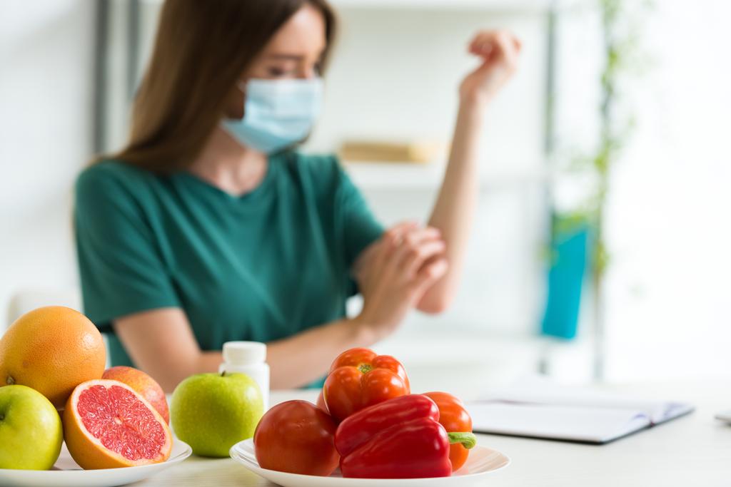szelektív középpontjában a fiatal nő az orvosi maszkot ül az asztalnál a gyümölcsök, zöldségek és tabletták és karmoló karját otthon - Fotó, kép