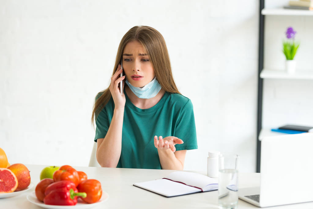 chica en máscara médica hablando en el teléfono inteligente en la mesa con frutas, verduras y pastillas en casa
 - Foto, Imagen