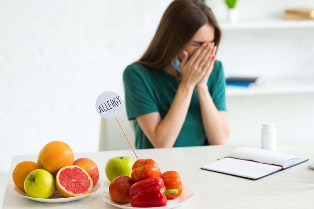 Žena se zdravotní maskou kýchání při posezení u stolu s ovocem, zeleninou a prášky - Fotografie, Obrázek