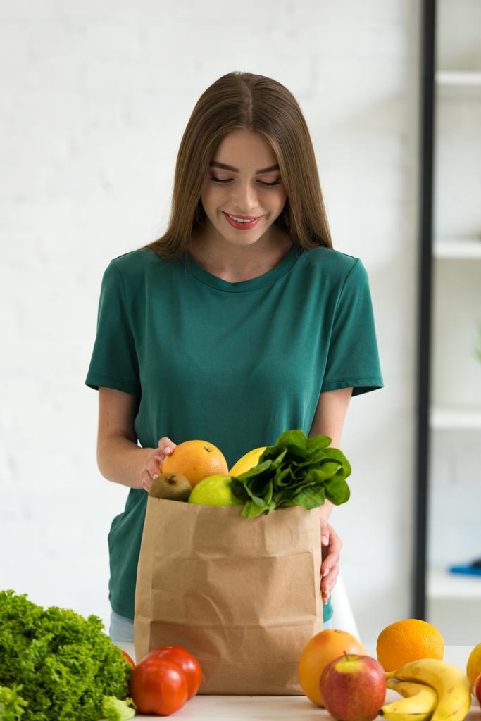sonriente joven mujer de pie cerca de bolsa de papel con verduras frescas y frutas en casa
 - Foto, Imagen