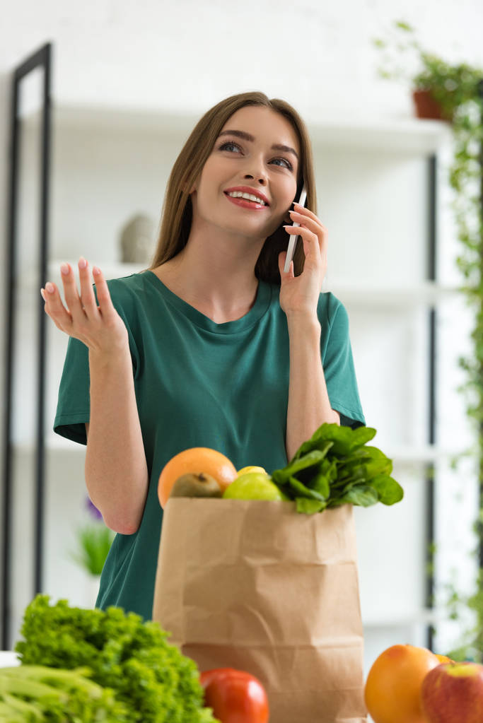 chica sonriente de pie cerca de la bolsa de papel con comida fresca y hablando en el teléfono inteligente en casa
 - Foto, imagen
