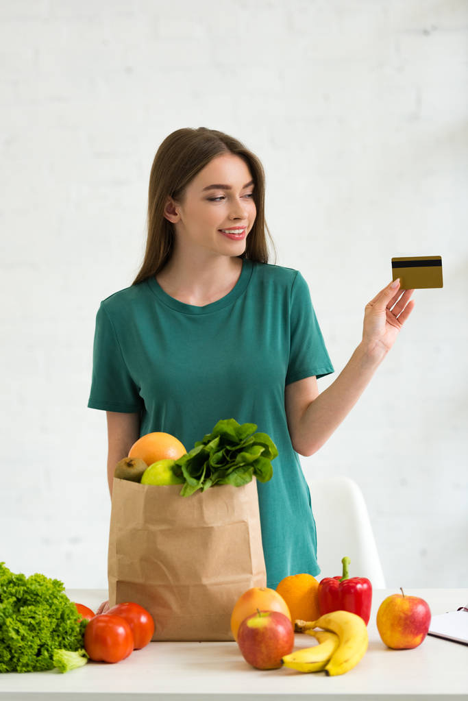 gülümseyen kız taze gıda ile kağıt torba yanında duran ve evde kredi kartı tutan - Fotoğraf, Görsel