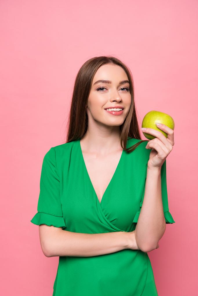 ελκυστική νεαρή γυναίκα που κρατά πράσινο μήλο και χαμογελά απομονωθεί σε ροζ - Φωτογραφία, εικόνα