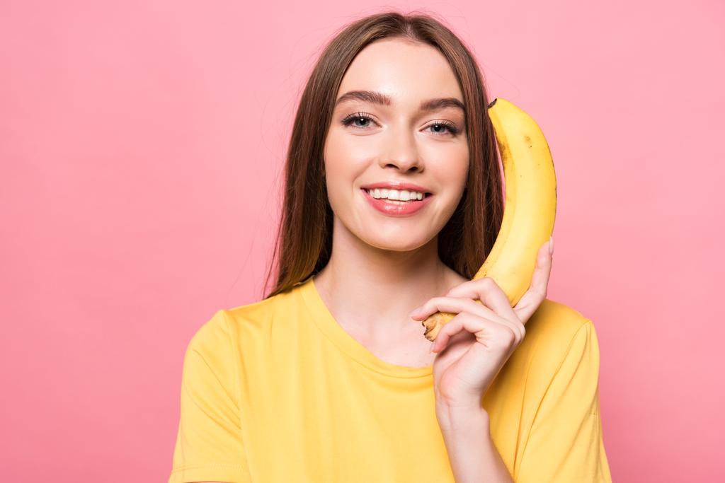 приваблива усміхнена дівчина тримає банан і дивиться на камеру ізольовано на рожевому
 - Фото, зображення