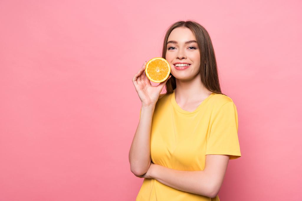 sonriente chica atractiva sosteniendo corte naranja y mirando a la cámara en rosa
 - Foto, Imagen