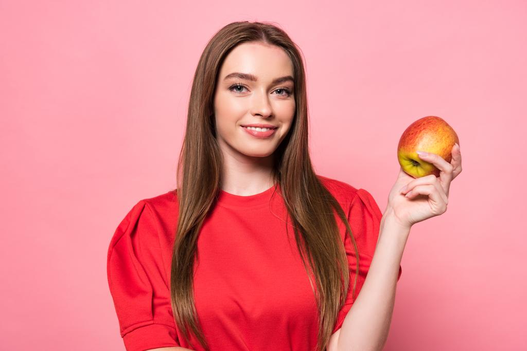 uśmiechnięta atrakcyjna dziewczyna trzyma jabłko i patrząc na kamerę na różowy - Zdjęcie, obraz
